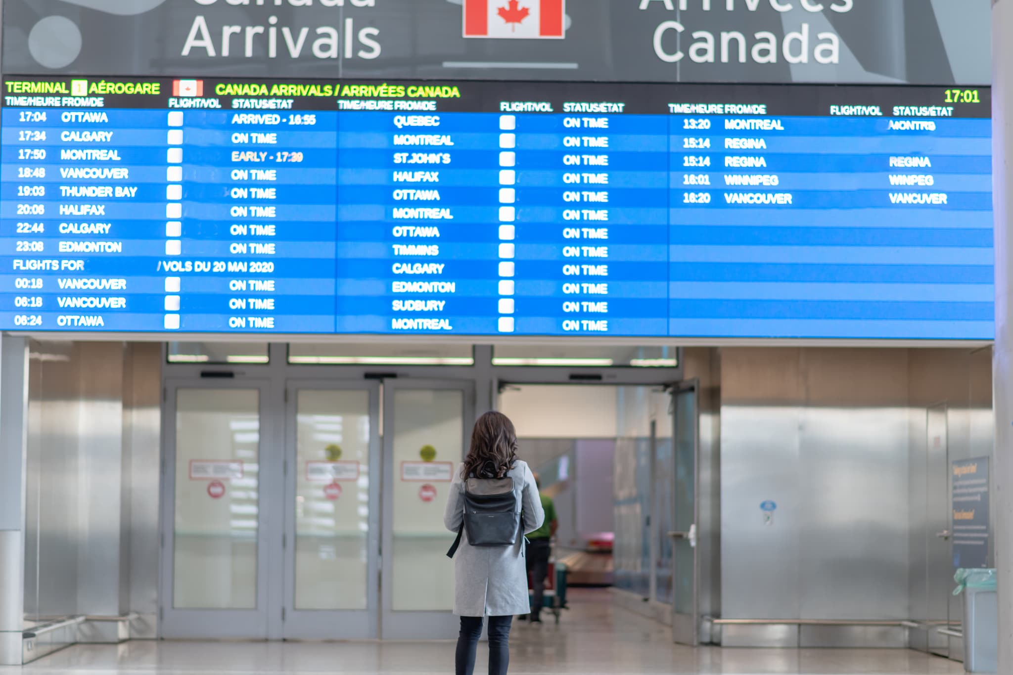 A young female (representing digital nomads in Canada) is standing in front of the arrival board waiting to be reunited with her husband. She is wearing a protective face mask. Travelling during the COVID-19 pandemic.