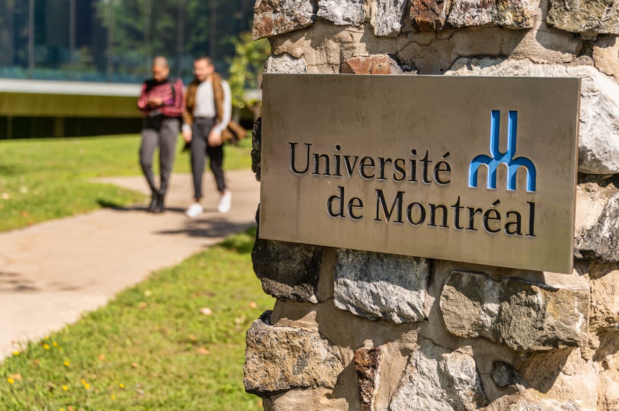 Université de Montréal sign mounted on a stone wall, with two people walking along a campus path in the background.