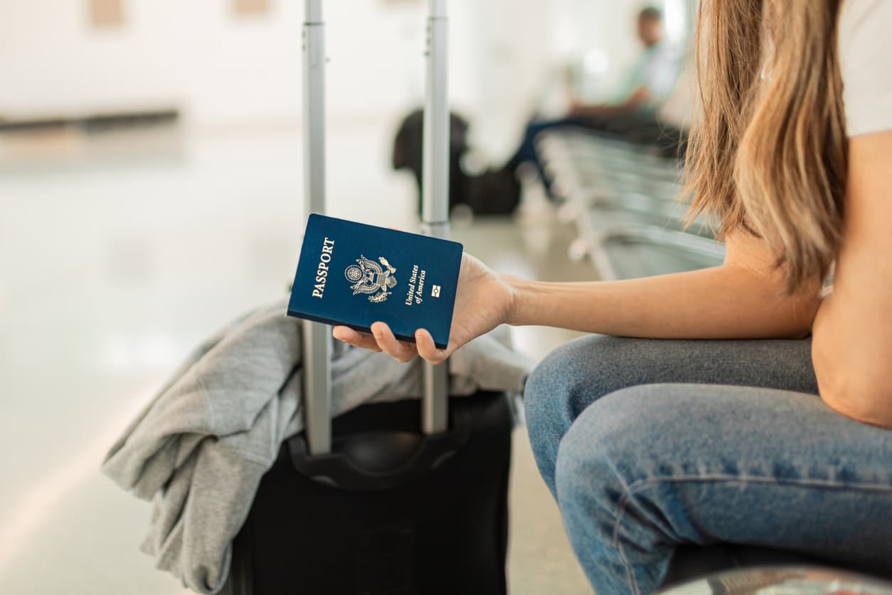 A traveler seated in an airport terminal holds a US passport beside a black rolling suitcase