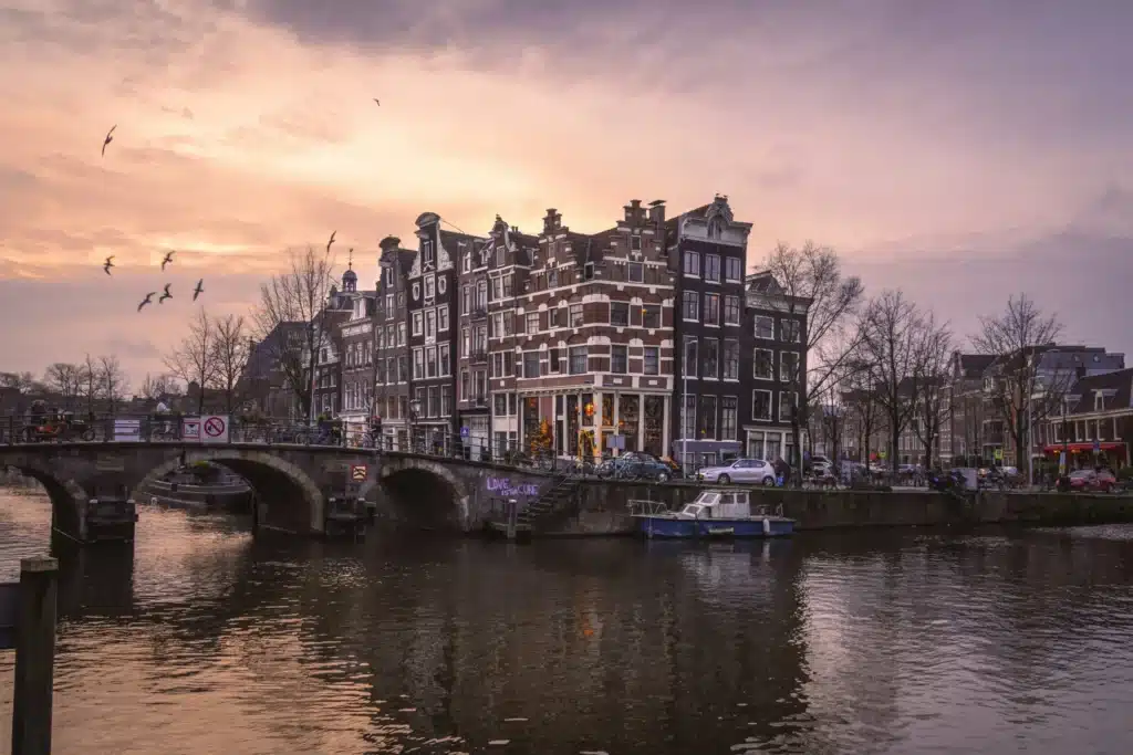 Transgender Healthcare in Europe: The Best Places for Gender-Affirming Care 2 A view of a canal in Amsterdam, Netherlands, at dusk. The image shows an arched stone bridge over the canal, and a cluster of tall, narrow historic buildings with dark brick facades is on the far bank.