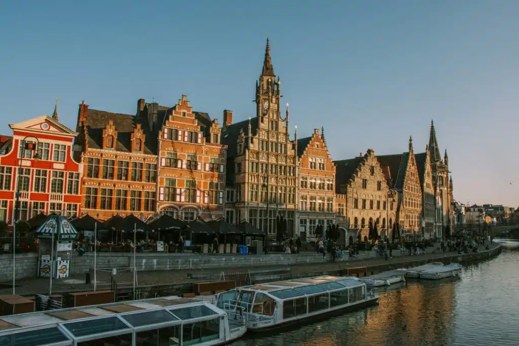 Transgender Healthcare in Europe: The Best Places for Gender-Affirming Care 1 A view of a historic canal in Ghent, Belgium, with a tour boat on the water. Along the canal, a row of old buildings with colorful brick facades and pointed roofs is lit by the warm light of the setting sun.