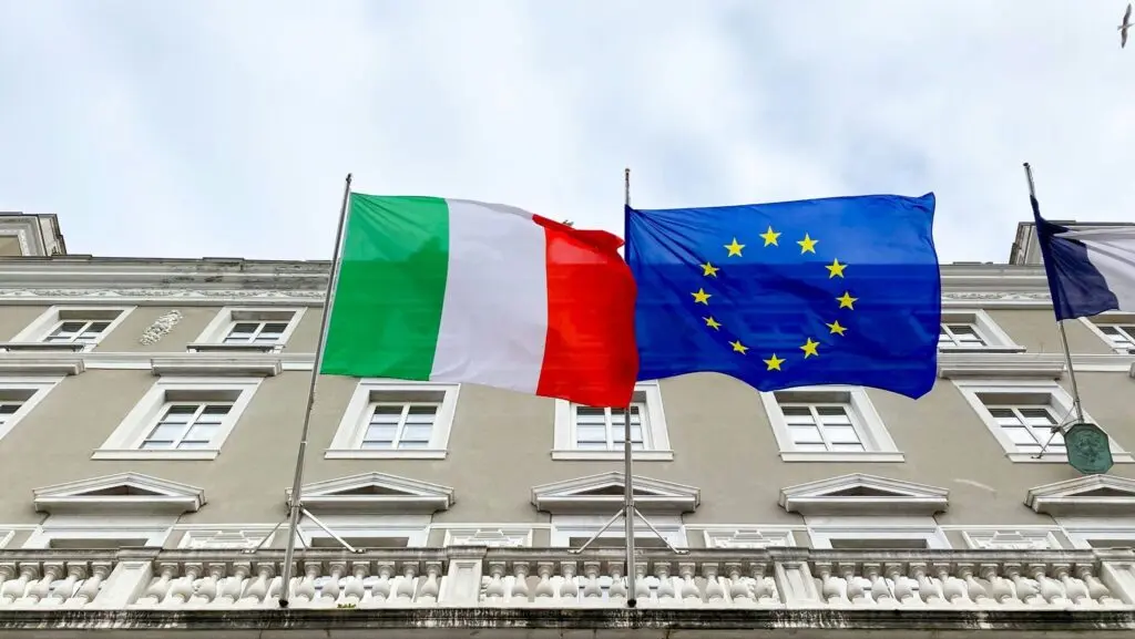 Italy High Court to Decide on Citizenship Reform in 2026 1 An Italian flag and an EU flag fly against a building backdrop; Italy's Supreme Court is set to rule on citizenship reform retroactivity in January 2026, affecting Italian citizenship by descent cases.