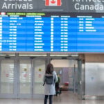 A young female (representing digital nomads in Canada) is standing in front of the arrival board waiting to be reunited with her husband. She is wearing a protective face mask. Travelling during the COVID-19 pandemic.