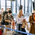 Passengers stand in a busy airport terminal, waiting in line for check-in and boarding while carrying luggage. Many people are engaged with their phones and preparing for their journey, similar to American moving back to the US for the first time after a long time living abroad.